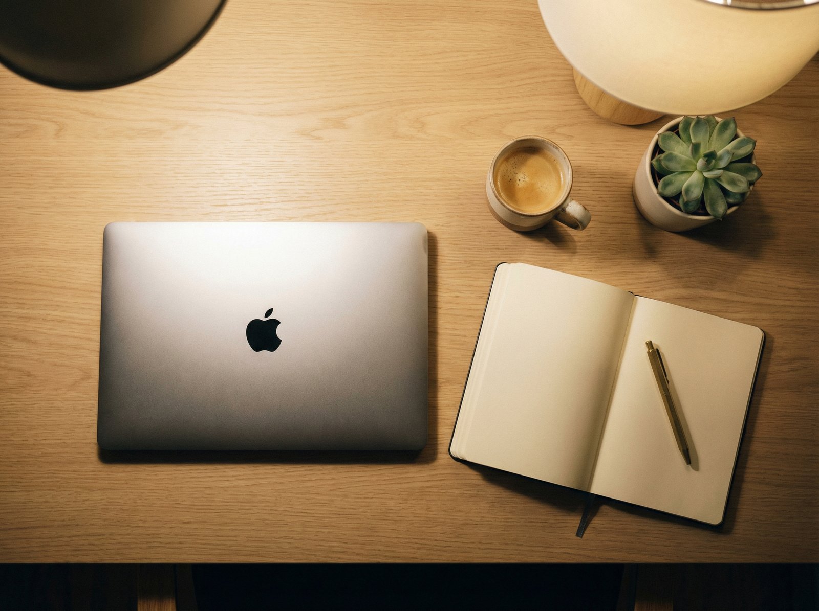 A brand new MacBook on a modern, clean desk with minimal accessories, an open notebook and a pen, warm, inviting lighting, top-down view, no visible text, aspect ratio 4:3