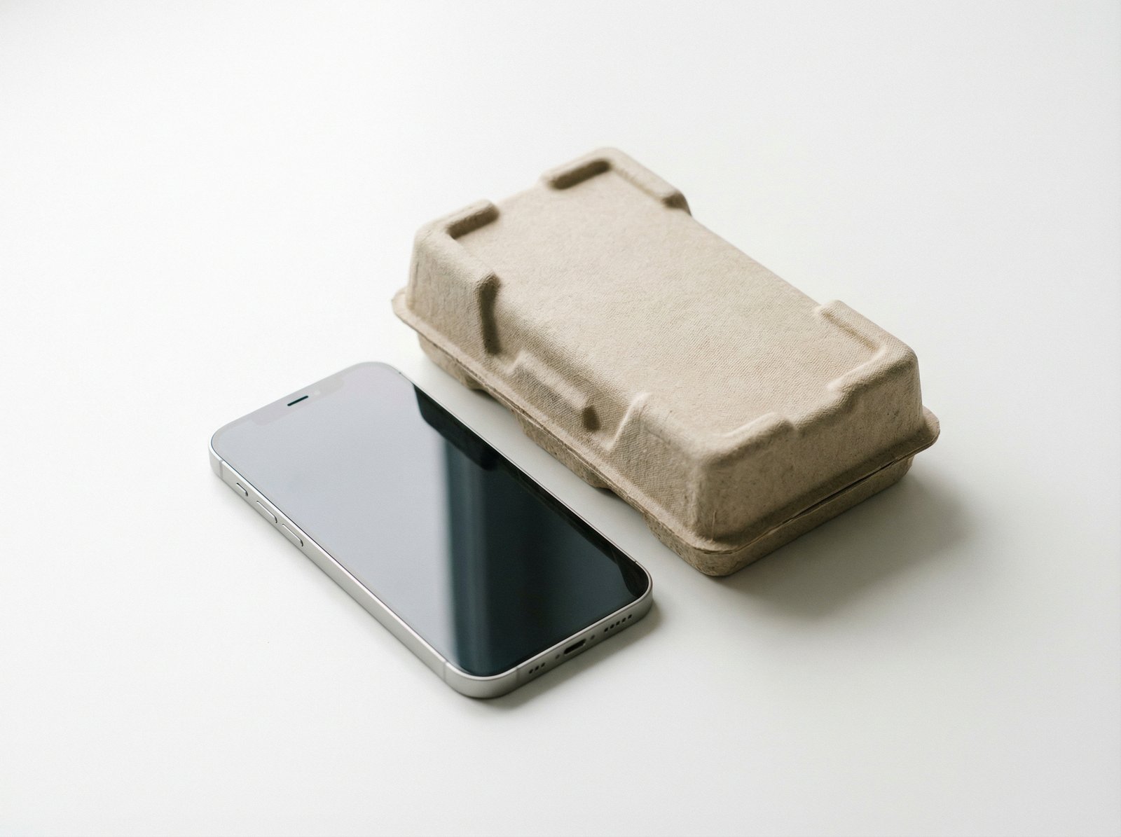 A professional studio shot of a sleek smartphone next to its eco-friendly paper packaging on a clean white surface with soft shadows, minimalist aesthetic, 4:3, no text