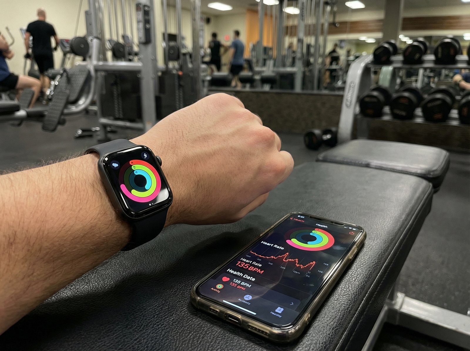 Close up shot of an Apple Watch and iPhone displaying fitness activity rings and heart rate data, gym background, realistic lighting, 4:3, no visible text