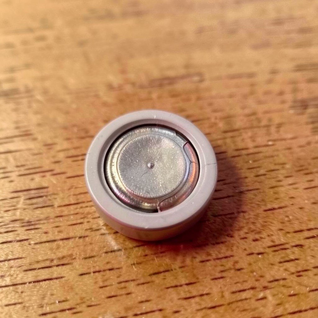 Macro view of an open round tracking tag showing a coin cell battery compartment and metal contacts on a wooden desk, no visible text, no logos, no watermark, aspect ratio 1:1